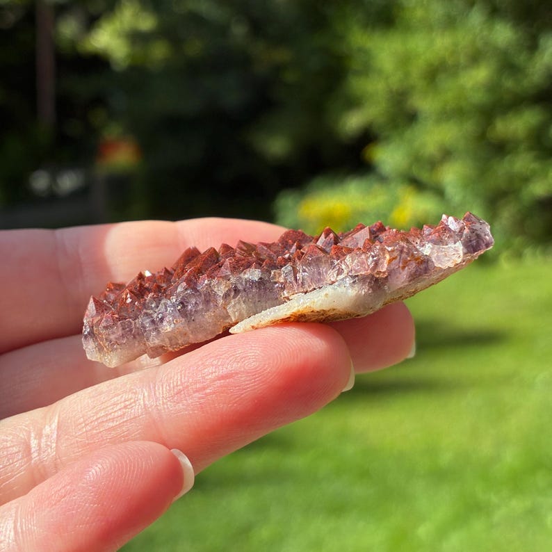 May include: A raw crystal cluster featuring a flat, light-colored base topped with numerous small, pointed formations. The crystals exhibit a gradient of deep reddish-brown at the tips transitioning into a translucent, pale purple hue at the base.