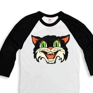 Op de afbeelding: Een wit en zwart baseballshirt met een zwarte kattenkop erop. De kat heeft groene ogen en een oranje mond. Het shirt heeft een zwarte kraag en zwarte mouwen.