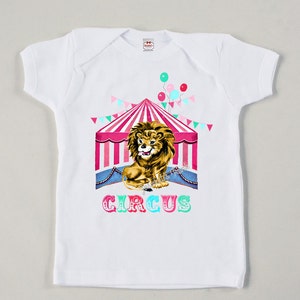 May include: White t-shirt featuring a colourful circus tent and a lion graphic. The text "CIRCUS" is printed below the tent in rainbow colours.