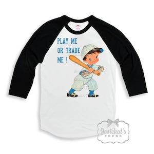 Könnte beinhalten: Ein weiß-schwarzes Baseball-Raglan-Shirt mit einer Cartoon-Illustration eines Jungen in Baseballuniform, der einen Schläger hält. Der Text "PLAY ME OR TRADE ME!" ist auf dem Shirt aufgedruckt.