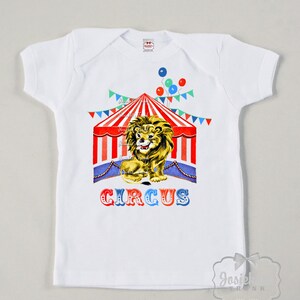 May include: White t-shirt featuring a red, white and blue circus tent graphic. A lion is sitting in front of the tent. The text "CIRCUS" is printed below the tent.