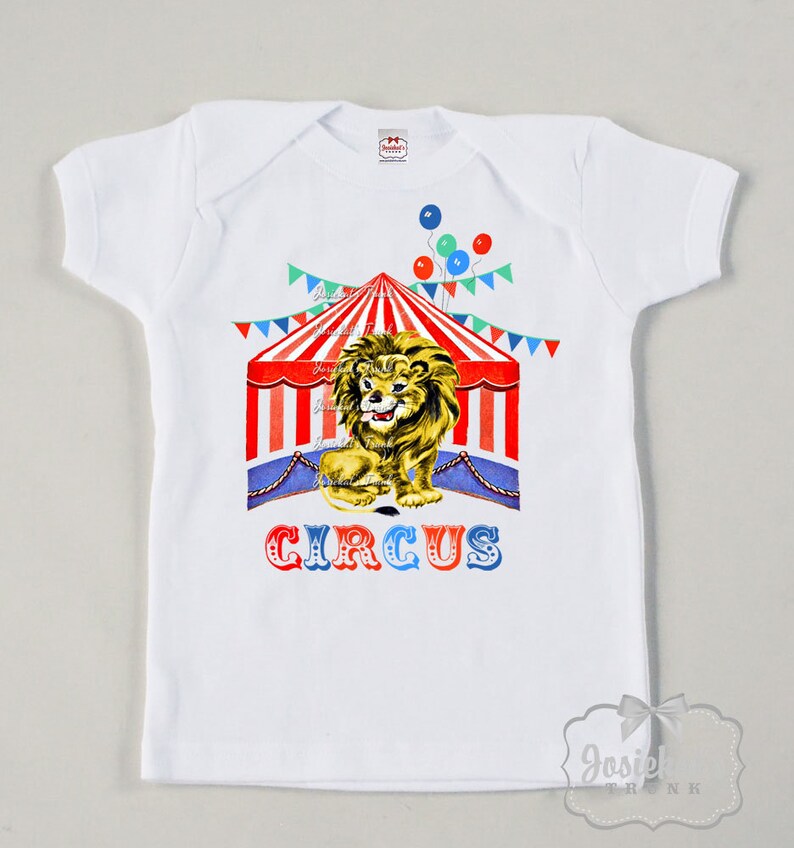 May include: White t-shirt featuring a red, white and blue circus tent graphic. A lion is sitting in front of the tent. The text "CIRCUS" is printed below the tent.