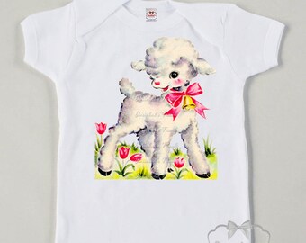 Peut inclure: Un body bébé blanc avec une illustration vintage d'un agneau blanc avec un nœud rose et une cloche, debout dans un champ d'herbe verte et de tulipes roses.