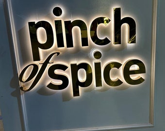 Può includere: Un cartello dorato con le parole "pinch of spice" in un carattere moderno. Il cartello è illuminato da luci bianche.