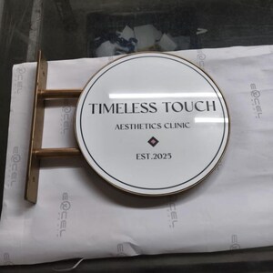 May include: A circular wall-mounted sign with a white face and a metallic gold frame. The text reads TIMELESS TOUCH AESTHETICS CLINIC EST.2025 with a small diamond icon. The sign is attached to a gold metal bracket with two horizontal support bars.
