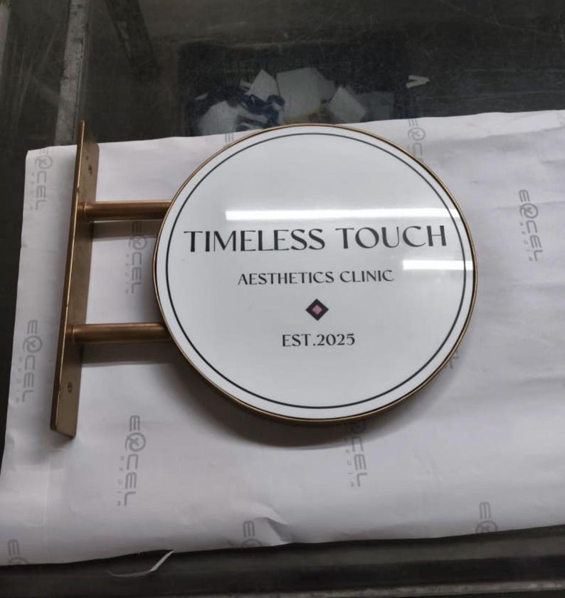 May include: A circular wall-mounted sign with a white face and a metallic gold frame. The text reads TIMELESS TOUCH AESTHETICS CLINIC EST.2025 with a small diamond icon. The sign is attached to a gold metal bracket with two horizontal support bars.