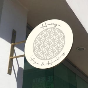 May include: A circular off-white sign mounted on a gold-toned metal bracket. The sign features a black line-art flower of life geometric pattern in the center, with the text "Haseya" above and "Yoga & Healing" below in a cursive script font.