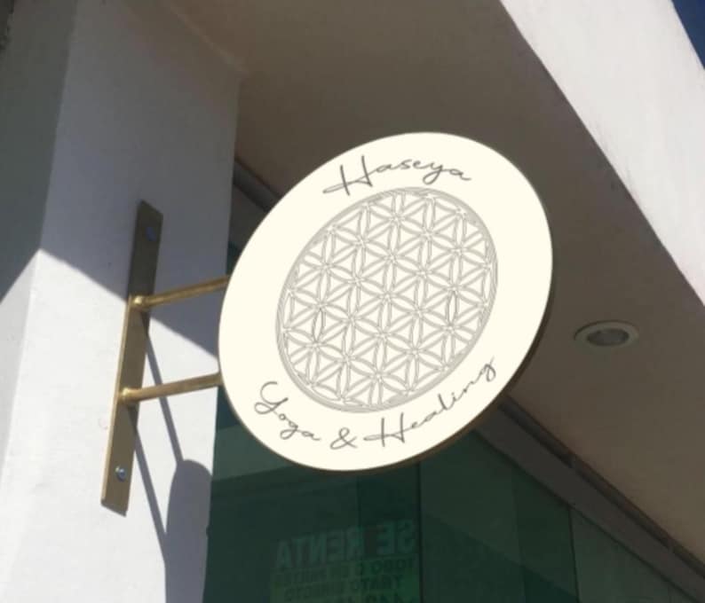 May include: A circular off-white sign mounted on a gold-toned metal bracket. The sign features a black line-art flower of life geometric pattern in the center, with the text "Haseya" above and "Yoga & Healing" below in a cursive script font.