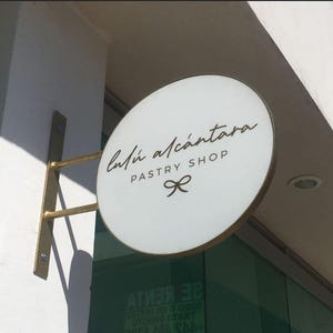 May include: A circular, white, double-sided projecting sign with a gold-toned metal frame and mounting bracket. The sign features the black text 'Lul&uacute; Alc&aacute;ntara' in a script font, 'PASTRY SHOP' in block letters, and a small bow icon below.