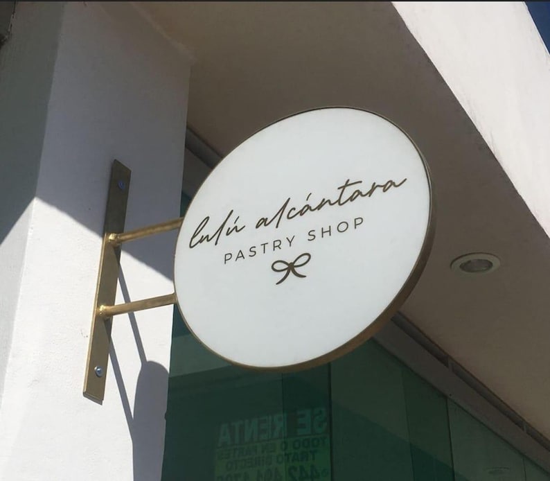 May include: A circular, white, double-sided projecting sign with a gold-toned metal frame and mounting bracket. The sign features the black text 'Lul&uacute; Alc&aacute;ntara' in a script font, 'PASTRY SHOP' in block letters, and a small bow icon below.