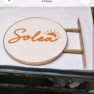 May include: A circular wall-mounted sign with a white face and a gold-toned metal frame. The word Solea is written in orange cursive script, accompanied by an orange graphic of a croissant with radiating lines above the letter a.