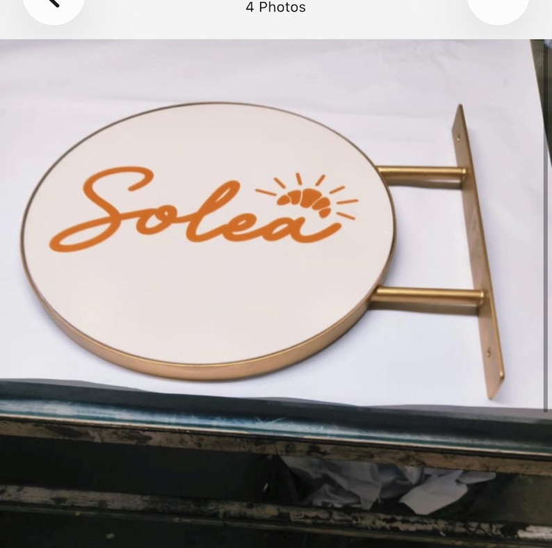 May include: A circular wall-mounted sign with a white face and a gold-toned metal frame. The word Solea is written in orange cursive script, accompanied by an orange graphic of a croissant with radiating lines above the letter a.