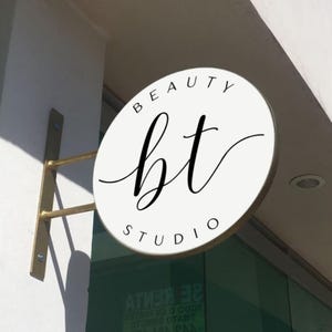 May include: A circular white sign with a gold metal mounting bracket attached to a wall. The sign features the black text BEAUTY STUDIO in a sans-serif font and the letters bt in a large, black, cursive script centered on the face of the sign.