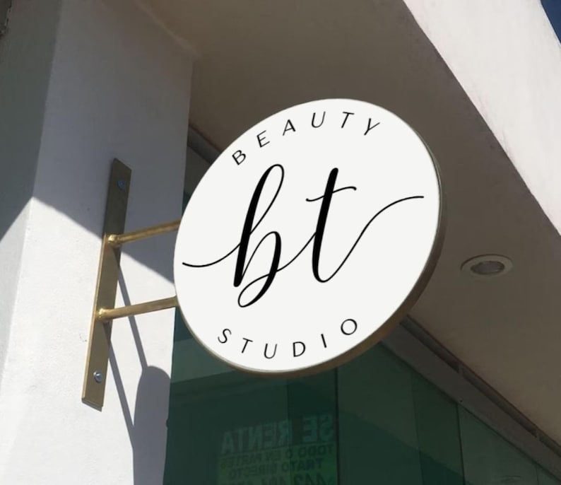 May include: A circular white sign with a gold metal mounting bracket attached to a wall. The sign features the black text BEAUTY STUDIO in a sans-serif font and the letters bt in a large, black, cursive script centered on the face of the sign.