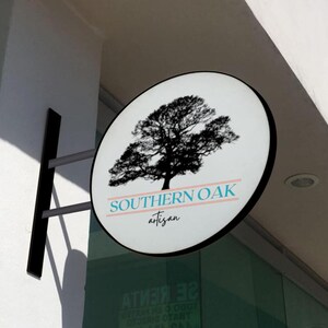 May include: A circular, wall-mounted sign featuring a black silhouette of an oak tree. Below the tree, the text reads SOUTHERN OAK in blue capital letters, flanked by two horizontal lines, with the word artisan in black script underneath.