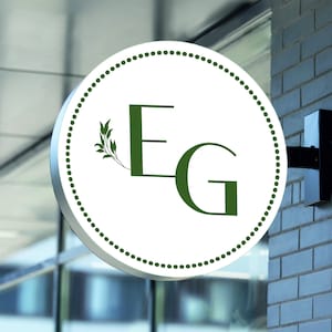May include: A white round sign with a green border and a green leafy design. The sign has the letters "EG" in green.