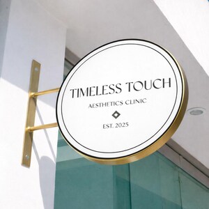 May include: A circular, double-sided sign with a gold-toned metal frame and mounting bracket. The white face features black text reading TIMELESS TOUCH AESTHETICS CLINIC, a small diamond-shaped icon, and the text EST. 2025 in a clean, serif font.
