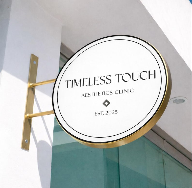 May include: A circular, double-sided sign with a gold-toned metal frame and mounting bracket. The white face features black text reading TIMELESS TOUCH AESTHETICS CLINIC, a small diamond-shaped icon, and the text EST. 2025 in a clean, serif font.