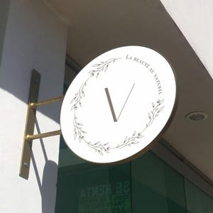 May include: A circular white wall-mounted sign with a gold-toned metal bracket. The sign features a central letter V surrounded by a delicate black line-art floral wreath and the text LA BEAUT&Eacute; AU NATUREL printed in a clean, black serif font.