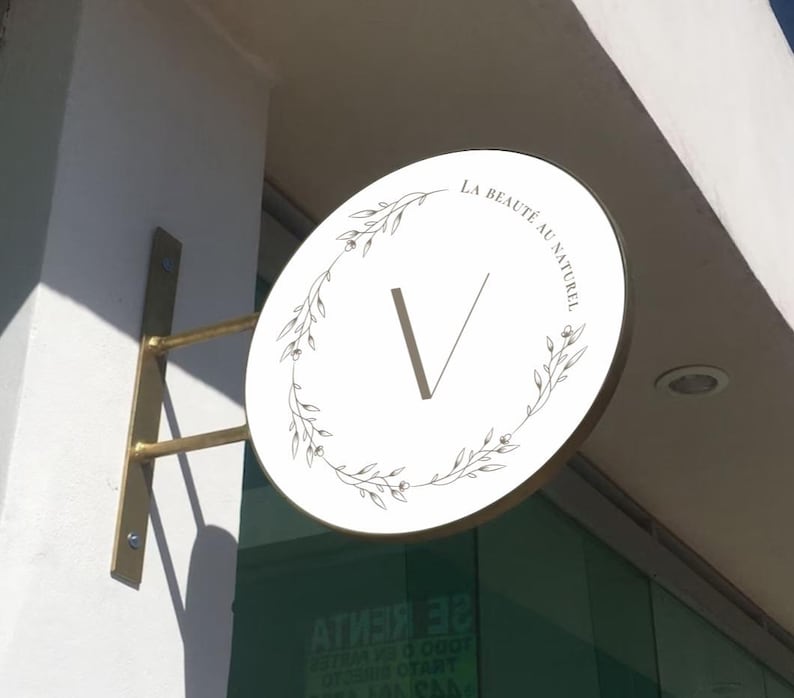 May include: A circular white wall-mounted sign with a gold-toned metal bracket. The sign features a central letter V surrounded by a delicate black line-art floral wreath and the text LA BEAUT&Eacute; AU NATUREL printed in a clean, black serif font.