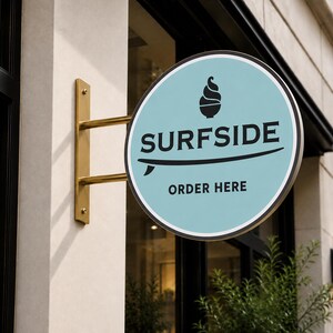 May include: A circular, light blue sign mounted on a gold-colored metal bracket. The sign features a black graphic of a soft-serve ice cream cone, the text SURFSIDE, a surfboard illustration, and the text ORDER HERE in black lettering.
