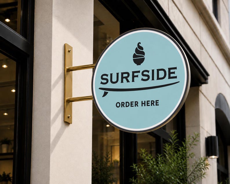 May include: A circular, light blue sign mounted on a gold-colored metal bracket. The sign features a black graphic of a soft-serve ice cream cone, the text SURFSIDE, a surfboard illustration, and the text ORDER HERE in black lettering.