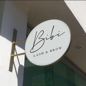 May include: A circular, white, double-sided projecting sign with a gold-colored metal bracket mounted to a wall. The sign features the black text 'Bibi' in a cursive script font, with 'LASH & BROW' printed in a smaller, capitalized sans-serif font.