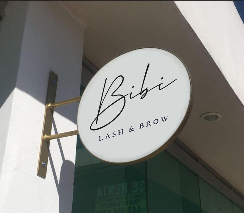 May include: A circular, white, double-sided projecting sign with a gold-colored metal bracket mounted to a wall. The sign features the black text 'Bibi' in a cursive script font, with 'LASH & BROW' printed in a smaller, capitalized sans-serif font.