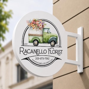 May include: A round white wall-mounted sign featuring a watercolor illustration of a green truck carrying a bouquet of colorful flowers. The text reads Racanello Florist, Est. 1936, 203-973-7562, and www.racanelloflorist.com in black lettering.