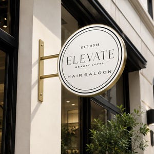 May include: A circular, double-sided metal sign with a gold-toned frame and mounting bracket. The white face features black circular borders and the text: EST. 2018, ELEVATE BEAUTY LOFTS, and HAIR SALOON, displayed in a clean, serif typeface.