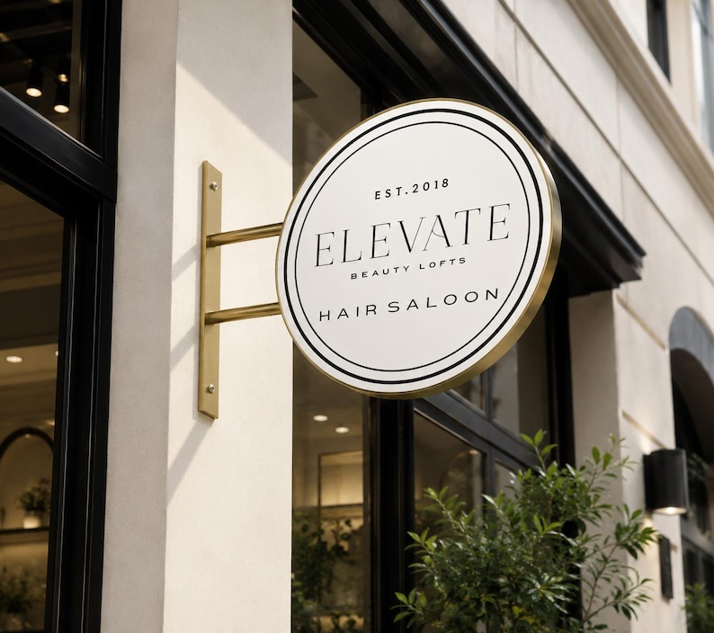 May include: A circular, double-sided metal sign with a gold-toned frame and mounting bracket. The white face features black circular borders and the text: EST. 2018, ELEVATE BEAUTY LOFTS, and HAIR SALOON, displayed in a clean, serif typeface.