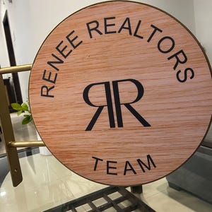 May include: A circular sign with a light wood grain finish, featuring the black text "RENEE REALTORS" at the top, "TEAM" at the bottom, and a central "RR" monogram. The sign is mounted on a gold-colored metal bracket with two horizontal arms.
