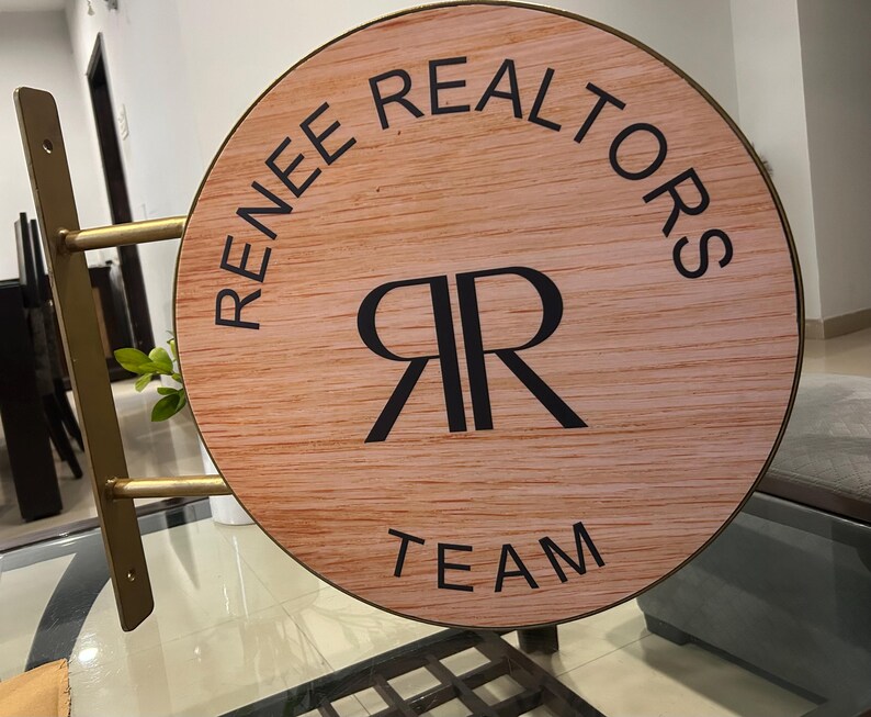 May include: A circular sign with a light wood grain finish, featuring the black text "RENEE REALTORS" at the top, "TEAM" at the bottom, and a central "RR" monogram. The sign is mounted on a gold-colored metal bracket with two horizontal arms.