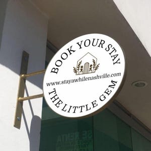 May include: A circular white sign mounted on a wall with a gold-toned metal bracket. The sign features black text reading BOOK YOUR STAY and THE LITTLE GEM, a line drawing of a house with plants, and the website www.stayawhilenashville.com.