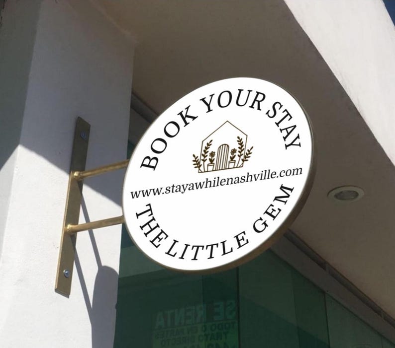 May include: A circular white sign mounted on a wall with a gold-toned metal bracket. The sign features black text reading BOOK YOUR STAY and THE LITTLE GEM, a line drawing of a house with plants, and the website www.stayawhilenashville.com.