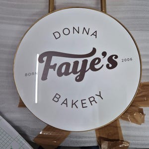 May include: A circular white sign with a gold metal frame and mounting bracket. The text reads DONNA in an arc, Faye's in large script, and BAKERY below. The words BORN and 2006 are printed on the left and right sides of the central script text.