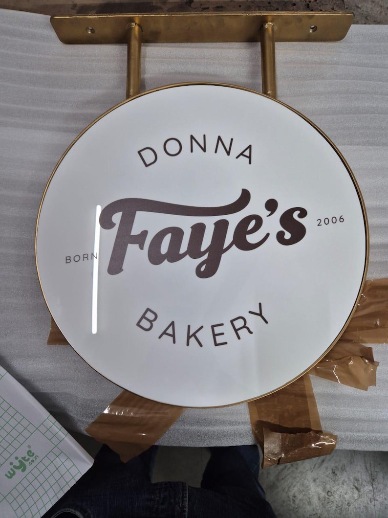 May include: A circular white sign with a gold metal frame and mounting bracket. The text reads DONNA in an arc, Faye's in large script, and BAKERY below. The words BORN and 2006 are printed on the left and right sides of the central script text.