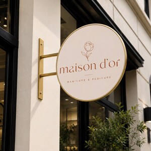 May include: A circular, cream-colored projecting sign with a gold metal frame and mounting bracket. The sign features a thin-line floral illustration above the text "maison d'or" and the words "MANICURE & PEDICURE" in a dark brown, sans-serif font.