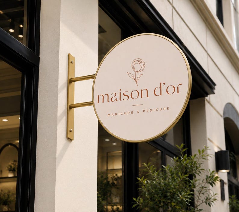May include: A circular, cream-colored projecting sign with a gold metal frame and mounting bracket. The sign features a thin-line floral illustration above the text "maison d'or" and the words "MANICURE & PEDICURE" in a dark brown, sans-serif font.