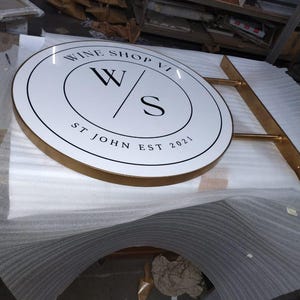 May include: A circular, white, double-sided sign with a gold-colored metal frame and mounting arm. The sign features black text reading WINE SHOP VI, a central W/S logo, and the text ST JOHN EST 2021, all enclosed within a thin black circular border.