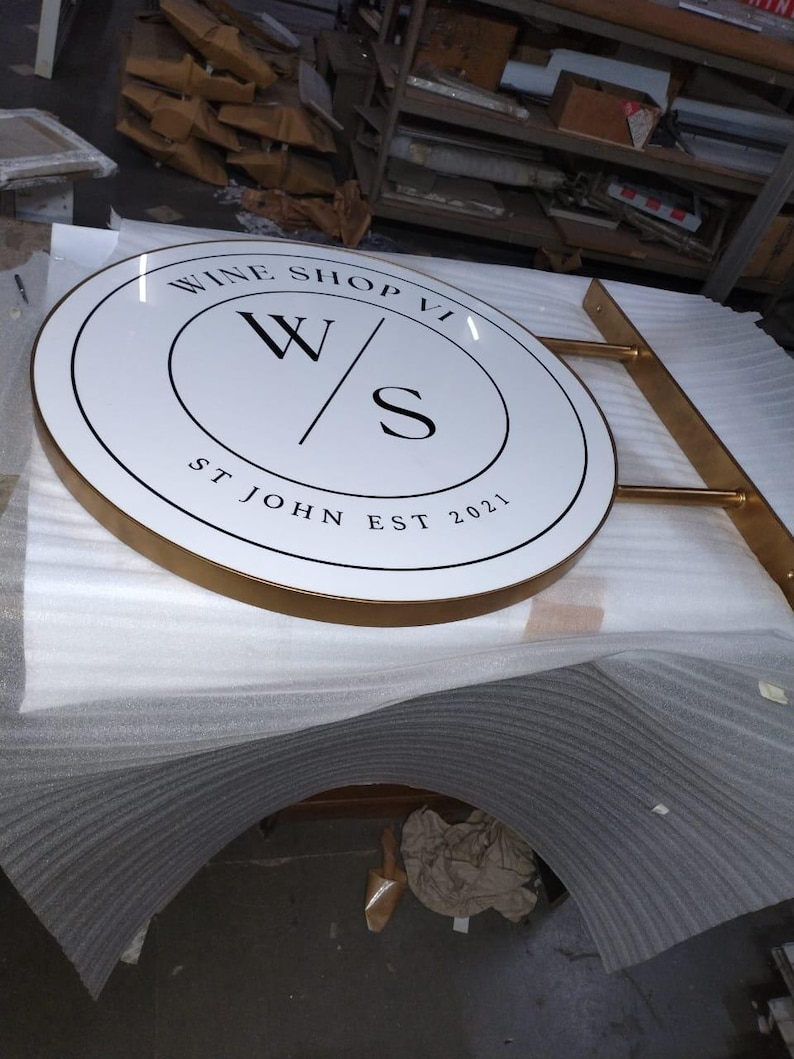 May include: A circular, white, double-sided sign with a gold-colored metal frame and mounting arm. The sign features black text reading WINE SHOP VI, a central W/S logo, and the text ST JOHN EST 2021, all enclosed within a thin black circular border.