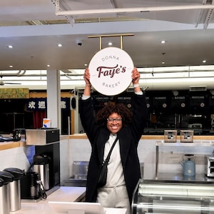 May include: A person holding a round sign for "Donna Faye's Bakery". The sign is white with a gold-colored frame and features the bakery's name in a burgundy script font. The bakery appears to be located in a commercial space.