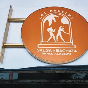 May include: A circular, orange-colored wall-mounted sign with a gold-toned metal bracket. The sign features white graphics of two dancing figures under an archway, with the text LOS ANGELES SALSA & BACHATA DANCE ACADEMY printed in white.