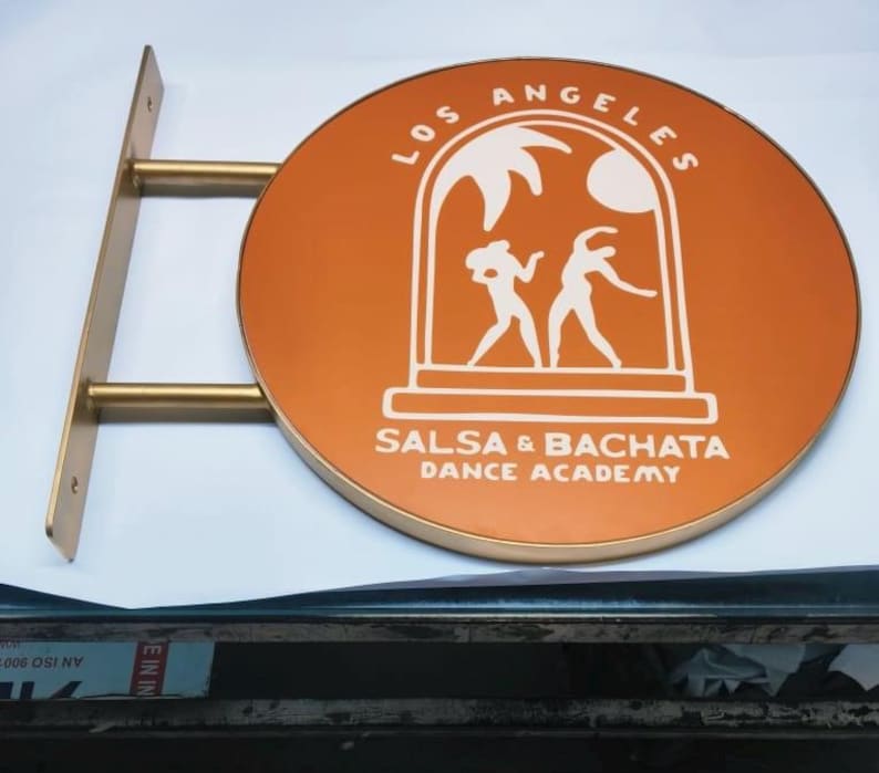 May include: A circular, orange-colored wall-mounted sign with a gold-toned metal bracket. The sign features white graphics of two dancing figures under an archway, with the text LOS ANGELES SALSA & BACHATA DANCE ACADEMY printed in white.