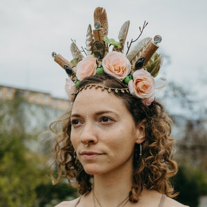 Könnte beinhalten: Eine Frau mit lockigem braunen Haar trägt eine Blumenkrone aus Federn, Kork und Kunstblumen. Die Krone ist mit kleinen goldenen Perlen und einem braunen Edelstein verziert. Sie trägt ein braunes Tanktop und eine Halskette mit einem großen braunen Steinanhänger.