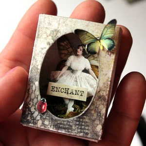 May include: A small, rectangular box with a vintage aesthetic. The box features a cutout window with a delicate, white-clad figure of a woman with wings. A butterfly with green and yellow wings is perched on the top of the box. The text "Enchant" is printed on a small banner below the figure. A red ladybug is visible on the bottom left corner of the box.