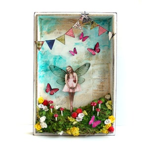 May include: A framed mixed media artwork featuring a fairy with pink wings and a pink dress, standing in a whimsical woodland setting with mushrooms, flowers, and butterflies. The background is a blue and green watercolour wash with a string of colourful flags.