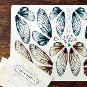 May include: A sheet of paper with a white background and a variety of insect wings printed on it. The wings are in shades of blue, green, and brown. The text "DOUBLE WINGS" is printed in black at the top of the sheet. The text "NICHOLA BATTILANA" is printed in black on the right side of the sheet. The text "PIXIEHILL.COM &copy; 2022" is printed in black below the text "NICHOLA BATTILANA".