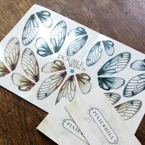 May include: A sheet of double wings, featuring a variety of insect wing designs in shades of blue and brown. The sheet is labelled "Double Wings" with a green four-leaf clover. Two business cards are visible in the foreground, one with the text "Pixie Hill" and the other with the text "Pixie".