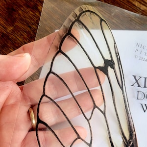 Puede incluir: Una lámina de plástico transparente con una imagen en blanco y negro de un ala de mariposa. El ala está detallada con venas y tiene una apariencia translúcida.